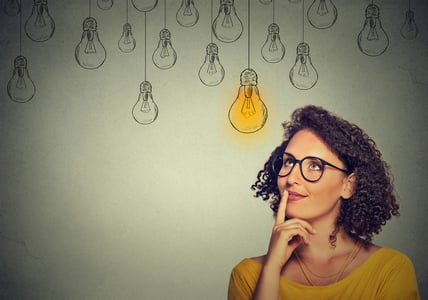 Thinking woman in glasses looking up with light idea bulb above head isolated on gray wall background-3