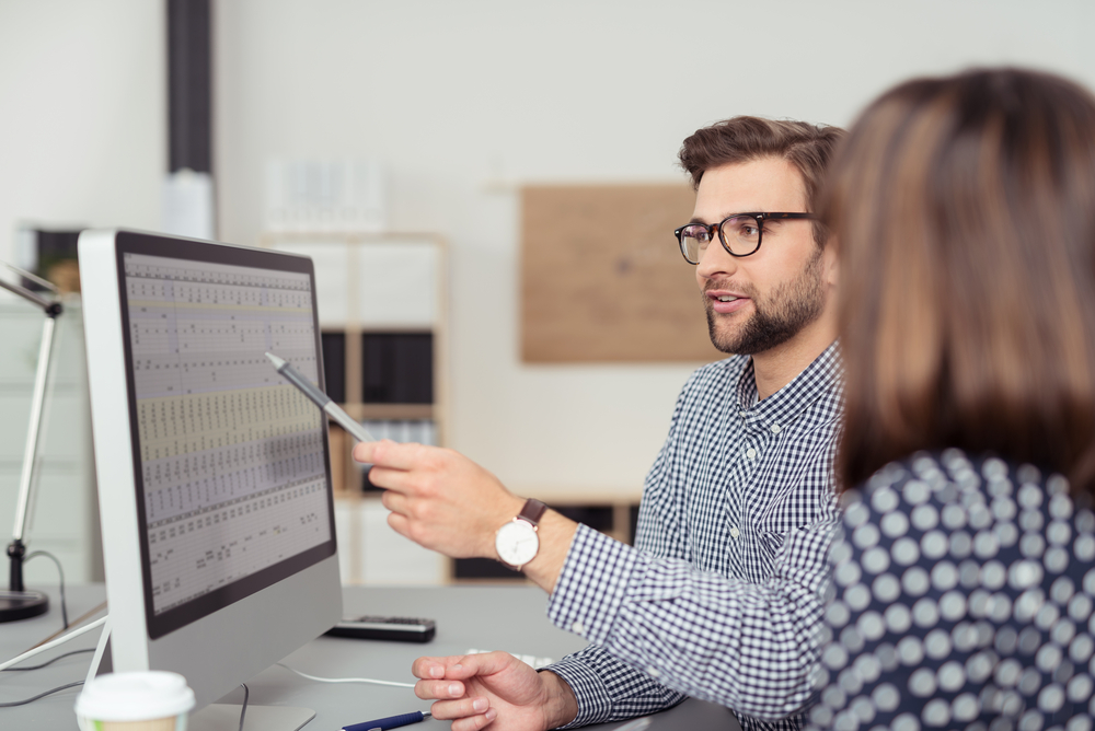 Proficient young male employee with eyeglasses and checkered shirt, explaining a business analysis displayed on the monitor of a desktop PC to his female colleague, in the interior of a modern office-Feb-29-2024-12-20-23-2239-AM