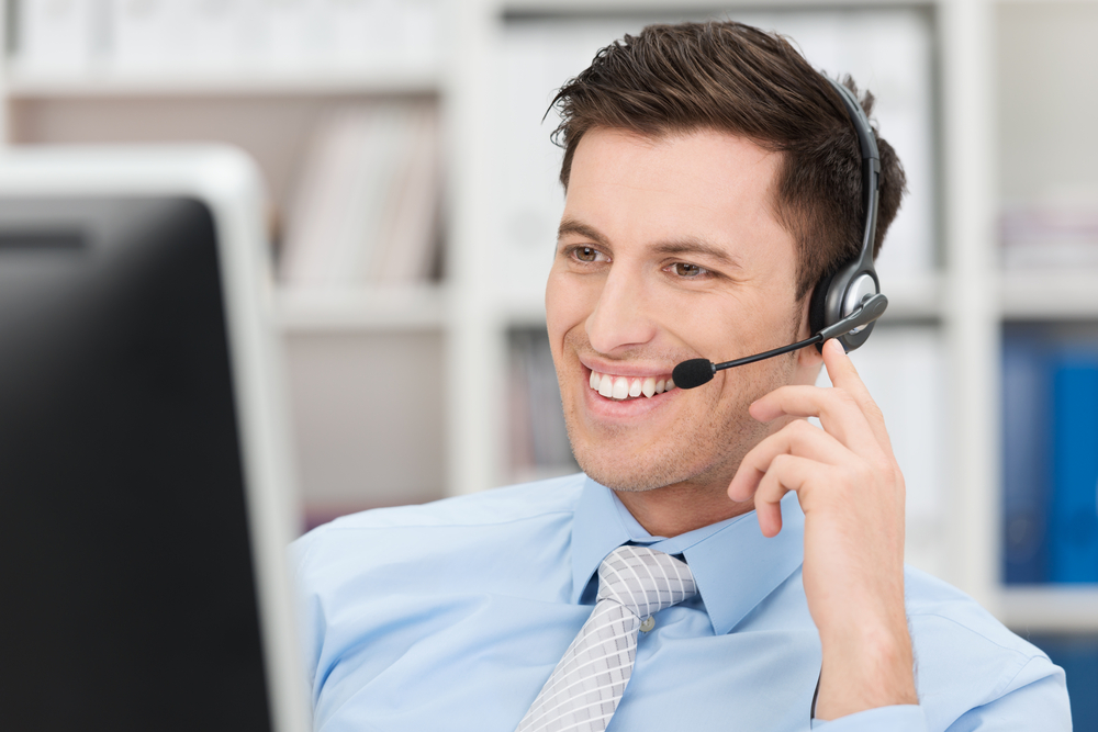 Smiling friendly handsome young male call centre operator or client services personnel beaming as he listens to a call and checks information on his computer monitor-1