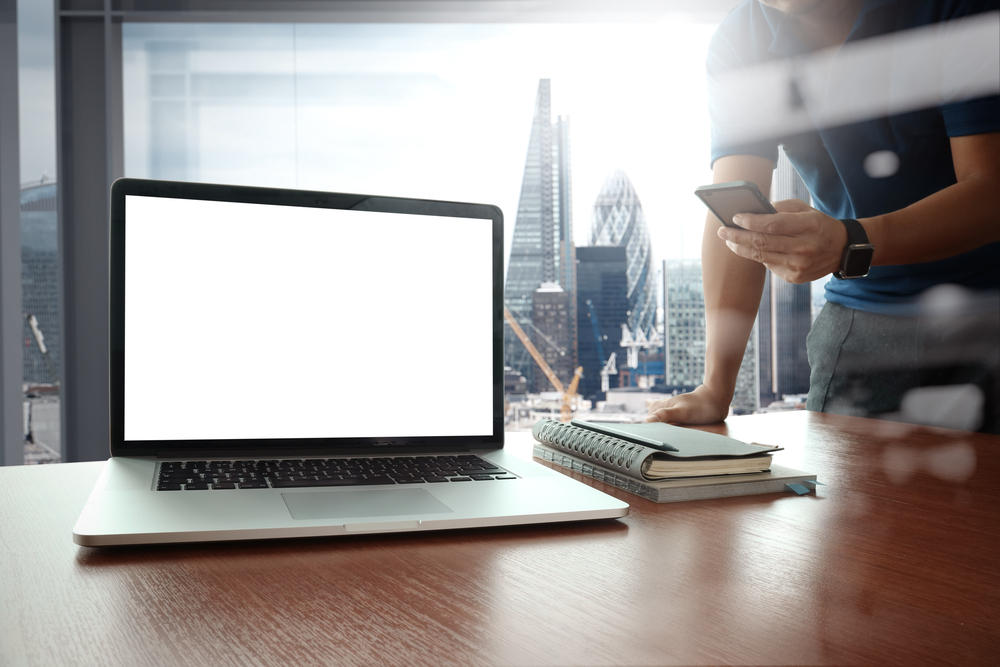 designer hand working and smart phone and blank screen laptop on wooden desk in office with london city background